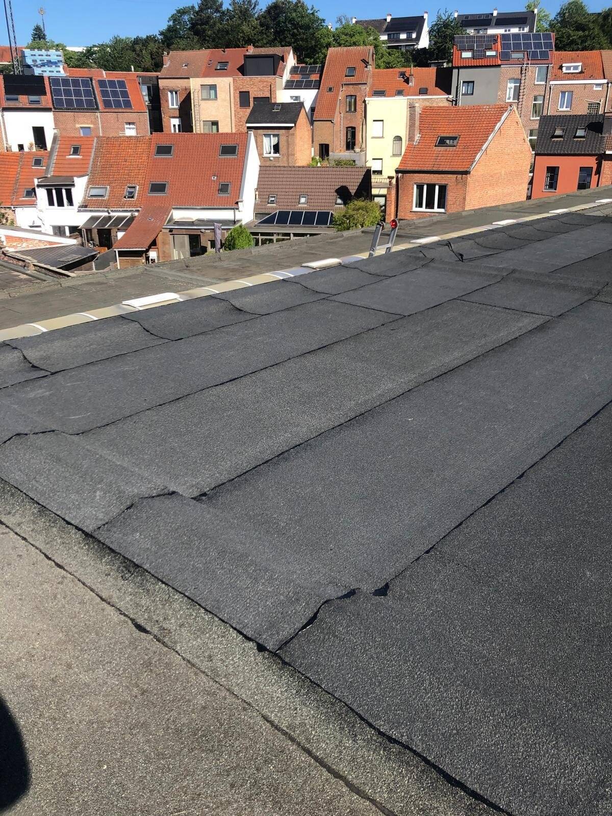 Gedetailleerd overzicht van een plat dak met verouderde bitumenbedekking en oneffenheden, met residentiële bebouwing op de achtergrond.